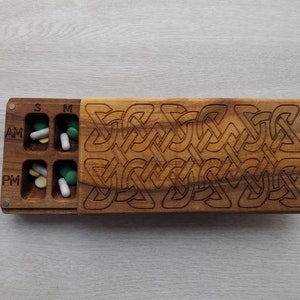 May include: A wooden pill box with a Celtic knot design. The box has two rows of compartments labeled "AM" and "PM" with "S", "M" and "S" above the compartments. The box is closed.