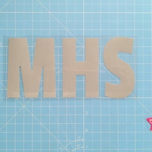 May include: A close-up of a gray vinyl sticker with the letters "MHS" cut out. The sticker is on a blue cutting mat with a white grid pattern.