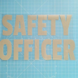 May include: A close-up of a safety officer sign made of silver vinyl letters on a clear background. The letters are cut out and placed on a blue cutting mat.