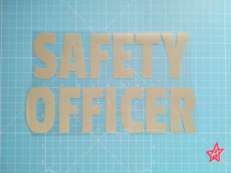 May include: A close-up of a safety officer sign made of silver vinyl letters on a clear background. The letters are cut out and placed on a blue cutting mat.