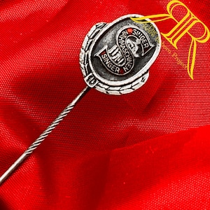 May include: A silver-coloured metal stick pin featuring an oval emblem with a textured border. The centre displays a black and red logo with the text SINGER and NAHMASCHINEN, centred around a stylised letter S and a small sewing machine icon.
