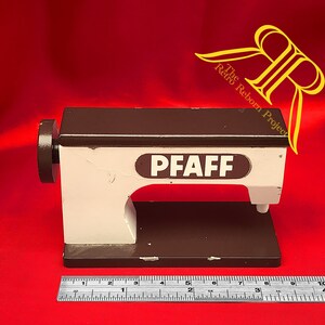 Könnte beinhalten: Ein Miniatur-Nähmaschinenmodell von Pfaff in Weiß und Dunkelbraun, ausgestattet mit einem seitlichen Handrad und einer Basis, die laut dem direkt vor dem Objekt platzierten Metalllineal etwa 24 cm lang ist.