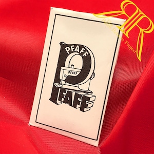 Op de afbeelding: Een rechthoekig wit stoffen label met een zwarte rand en een zwart grafisch logo. Het logo toont een grote letter P met daarin een illustratie van een naaimachine en het woord PFAFF boven en onder de machineafbeelding.