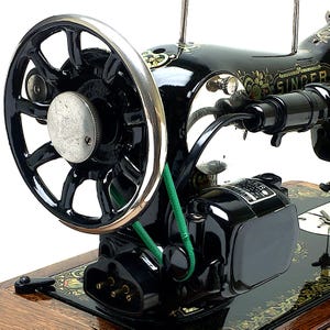 Pode incluir: Uma máquina de costura Singer preta vintage com um volante de borda prateada, uma correia de transmissão verde e um motor elétrico. O corpo da máquina é decorado com motivos florais dourados e inclui uma base de madeira.