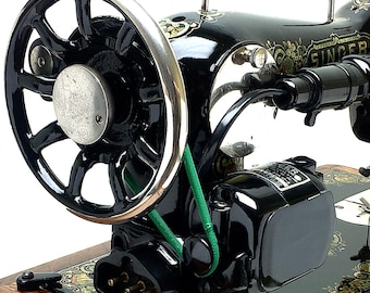 May include: A vintage black Singer sewing machine featuring a prominent silver-rimmed handwheel, a green drive belt, and an attached electric motor. The machine body is decorated with gold floral decals and includes a wooden base plate.