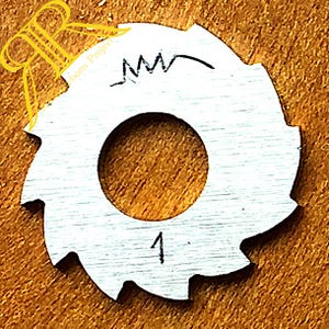 May include: A circular silver-colored metal disc with a serrated outer edge and a large central hole. The surface features a hand-drawn black ink symbol resembling a jagged line above the hole and the number 1 printed below the hole.