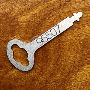 Pode incluir: Uma chave de corda de metal prateado, plana, com uma cabeça arredondada com um recorte central e um eixo longo e estreito. O número 96507 está gravado claramente a preto na superfície do eixo, que termina numa pequena ponta entalhada.