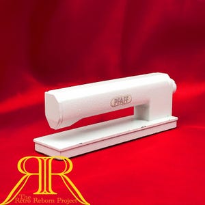 May include: A white metal sewing machine attachment with a rectangular base and a horizontal arm. The brand name PFAFF is printed in black on the side of the arm. The object has a smooth, painted finish and a sturdy, industrial construction.