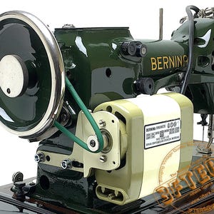Può includere: Una macchina da cucire Bernina verde scuro con un volantino cromato e un motore verde chiaro fissato sul lato. Il motore presenta un'etichetta bianca con specifiche tecniche e il marchio Bernina stampato in oro sul corpo della macchina.