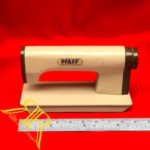 Op de afbeelding: Een crèmekleurige metalen naaimachine-arm met bruine accenten en de merknaam PFAFF in het zwart gedrukt. Een metalen liniaal staat ervoor en laat zien dat de basis ongeveer 23 cm lang is van de ene naar de andere kant.