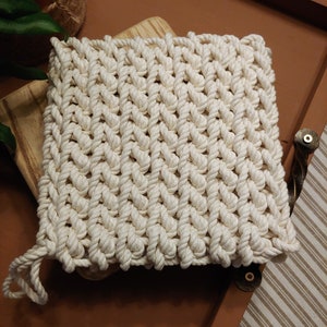 May include: A white crocheted pot holder with a woven pattern. The pot holder is made of thick cotton rope and has a square shape.