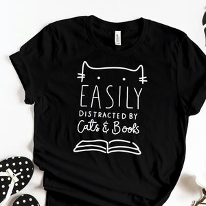 May include: Black t-shirt with white text that reads "Easily Distracted by Cats & Books". The text is stylized with a cat head and an open book.
