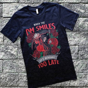 May include: A dark blue t-shirt with a graphic of a grim reaper holding a dice and a scythe. The text on the shirt reads "When the DM Smiles It's Already Too Late".