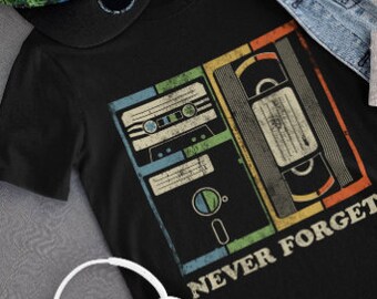 May include: Black t-shirt with a vintage-style graphic of a cassette tape, floppy disk, and VHS tape. The graphic is surrounded by a rainbow-colored border. The text "Never Forget" is printed below the graphic.