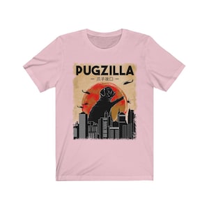 May include: A pink t-shirt with a black graphic of a pug standing on a city skyline. The graphic says "Pugzilla -  パグジラ - "