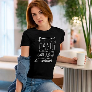 May include: Black t-shirt with white text that reads "Easily Distracted by Cats & Books". The text is accompanied by a simple line drawing of a cat's head with whiskers.