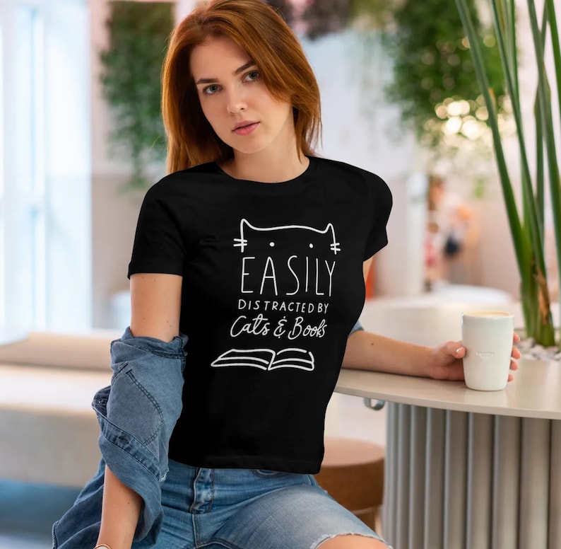 May include: Black t-shirt with white text that reads "Easily Distracted by Cats & Books". The text is accompanied by a simple line drawing of a cat's head with whiskers.