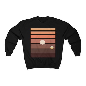 Peut inclure: Sweat-shirt noir avec un graphique représentant un coucher de soleil avec deux planètes. Le graphique est dans des tons de brun, orange et jaune.
