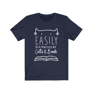 May include: Navy blue t-shirt with a white outline of a cat and an open book. The text "Easily Distracted by Cats & Books" is printed on the shirt.