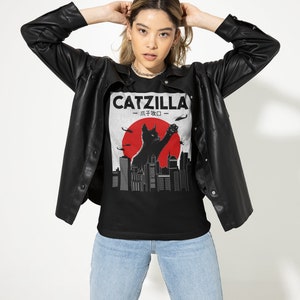May include: A black t-shirt with a graphic print of a black cat standing on a city skyline with the text "CATZILLA - 小子 奴 -" in white.