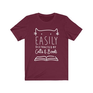 May include: A maroon t-shirt with a white graphic design that says "Easily Distracted by Cats & Books". The graphic also includes a stylized cat and an open book.