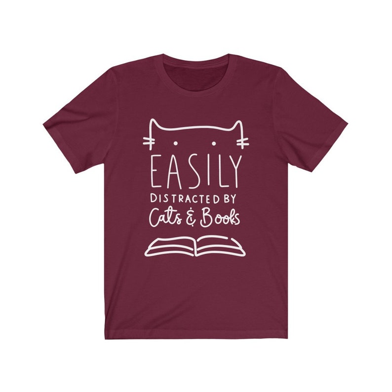 May include: A maroon t-shirt with a white graphic design that says "Easily Distracted by Cats & Books". The graphic also includes a stylized cat and an open book.