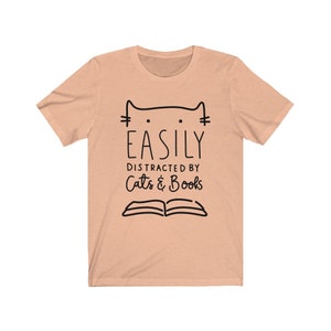 May include: A peach-colored t-shirt with a black cat illustration and the text "EASILY DISTRACTED BY Cats & Books".