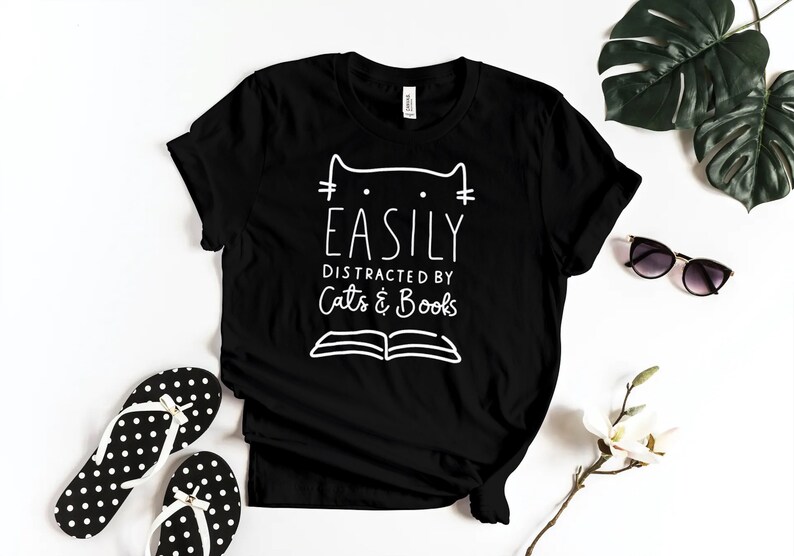 May include: Black t-shirt with white text that reads "Easily Distracted by Cats & Books". The text is stylized with a cat head and an open book.