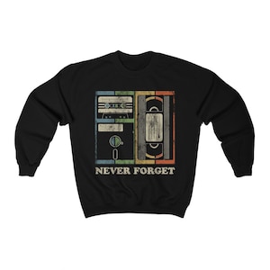 May include: Black sweatshirt with a vintage-style graphic of a cassette tape, floppy disk, and VHS tape. The graphic is surrounded by a colorful border and the text "NEVER FORGET".
