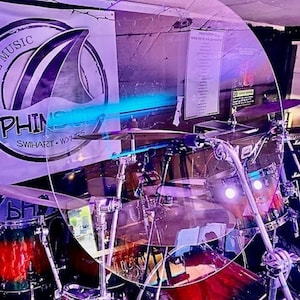 May include: A clear plastic drum shield with a purple and blue background. The shield is protecting a drum kit from the sound of other instruments.