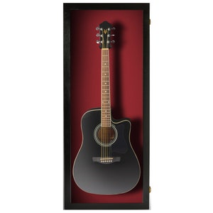 May include: Acoustic guitar displayed in a black framed shadow box. The guitar is black with a natural wood neck and gold tuning pegs. The background is a deep red color.