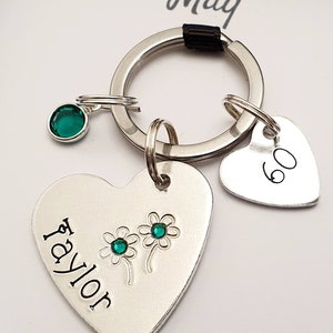 May include: A silver heart-shaped keychain with the name "Taylor" engraved on it. Two green gemstones are embedded in the heart, representing flowers. A smaller silver heart charm with the number "60" engraved on it is attached to the keychain. The keychain is attached to a silver ring.