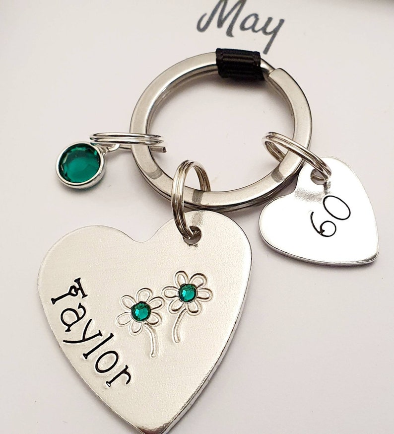 May include: A silver heart-shaped keychain with the name "Taylor" engraved on it. Two green gemstones are embedded in the heart, representing flowers. A smaller silver heart charm with the number "60" engraved on it is attached to the keychain. The keychain is attached to a silver ring.