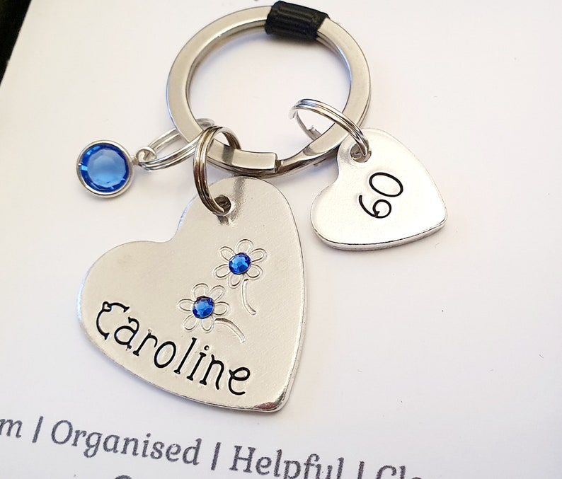 May include: Silver heart-shaped keychain with the name Caroline engraved on it, a blue rhinestone charm, and a smaller heart-shaped charm with the number 60 engraved on it.