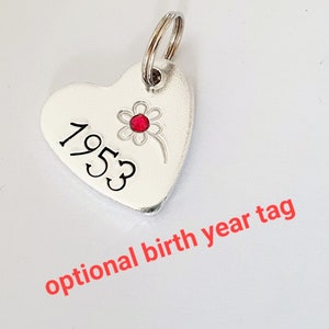 May include: Silver heart-shaped tag with a red rhinestone flower and the year 1953 engraved on it.  Optional birth year tag.
