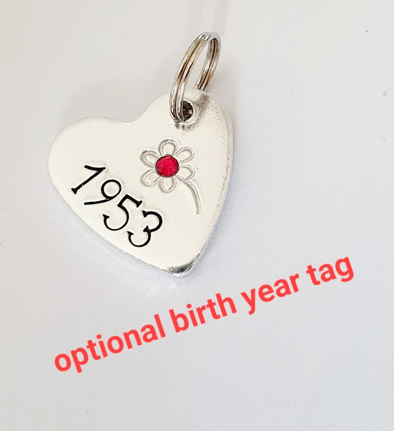 May include: Silver heart-shaped tag with a red rhinestone flower and the year 1953 engraved on it.  Optional birth year tag.
