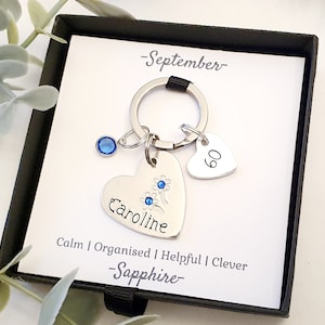 May include: A silver heart-shaped keychain with the name "Caroline" engraved on it. The keychain also features a small blue crystal charm and a heart-shaped charm with the number "90" on it. The keychain is presented in a black gift box with a white card that reads "September - Sapphire - Calm | Organised | Helpful | Clever".