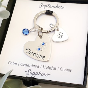 May include: A silver heart-shaped keychain with the name "Caroline" engraved on it. The keychain also features a small blue gemstone and a smaller heart charm with the number "60" engraved on it. The keychain is displayed in a black gift box with a white card that reads "September - Sapphire - Calm | Organised | Helpful | Clever".