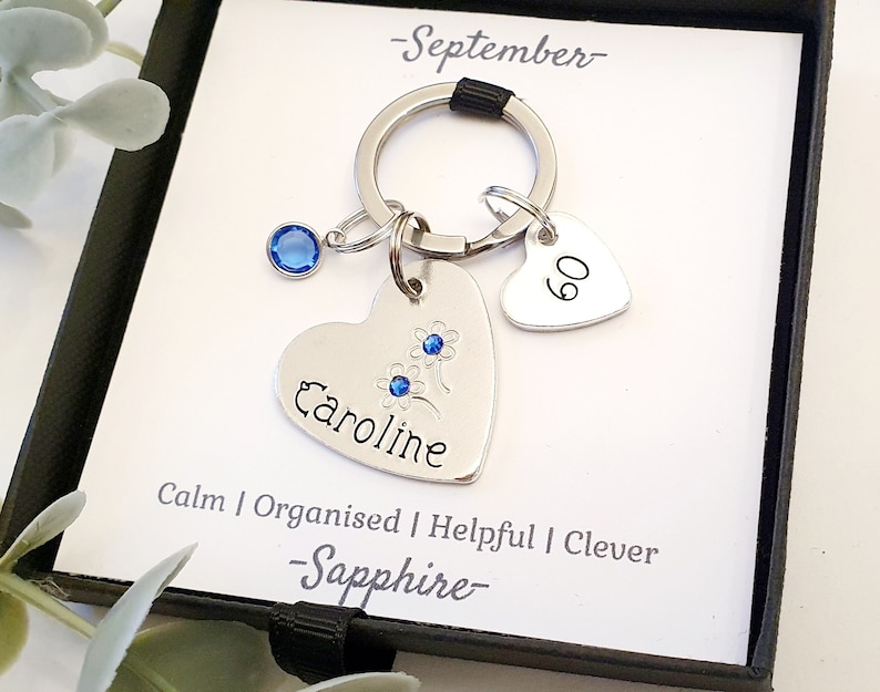 May include: A silver heart-shaped keychain with the name "Caroline" engraved on it. The keychain also features a small blue gemstone and a smaller heart charm with the number "60" engraved on it. The keychain is displayed in a black gift box with a white card that reads "September - Sapphire - Calm | Organised | Helpful | Clever".
