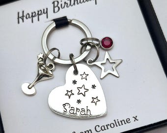 May include: Silver heart-shaped keychain with a star design and the name "Sarah" engraved on it. The keychain also features a small martini glass charm, a star charm, and a red rhinestone. The keychain is displayed in a black gift box with a white card that reads "Happy Birthday" and "Love from Caroline x".