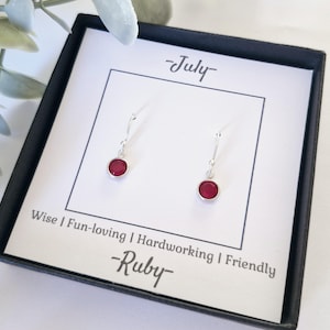 May include: A small white box containing a pair of red ruby gemstone earrings. The box is labelled with the text "July - Ruby" and "Wise | Fun-loving | Hardworking | Friendly".