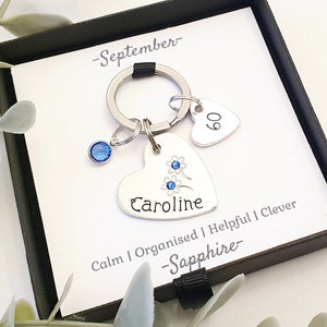 May include: A silver heart-shaped keychain with a blue crystal charm, a small heart charm with the number 60 on it, and the name "Caroline" engraved on the larger heart. The keychain also has the words "Calm | Organised | Helpful | Clever - Sapphire - September -" engraved on it.