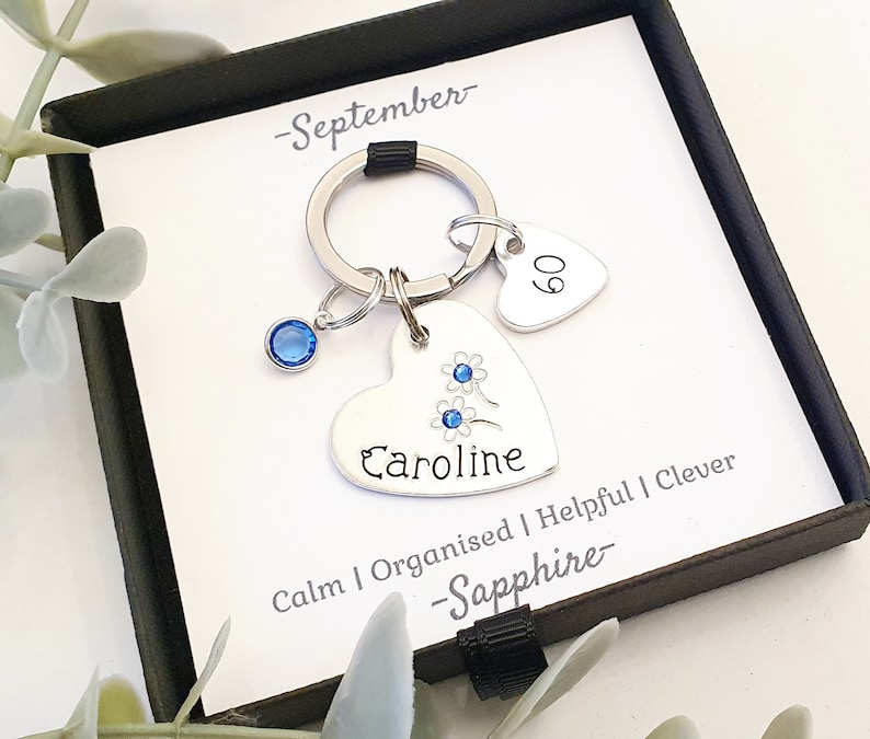 May include: A silver heart-shaped keychain with a blue crystal charm, a small heart charm with the number 60 on it, and the name "Caroline" engraved on the larger heart. The keychain also has the words "Calm | Organised | Helpful | Clever - Sapphire - September -" engraved on it.