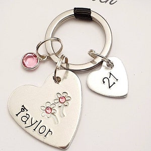 May include: Silver heart-shaped keychain with the name "Taylor" engraved on it, two pink flowers, and a pink rhinestone. A smaller silver heart-shaped charm with the number "21" engraved on it is attached to the keychain.