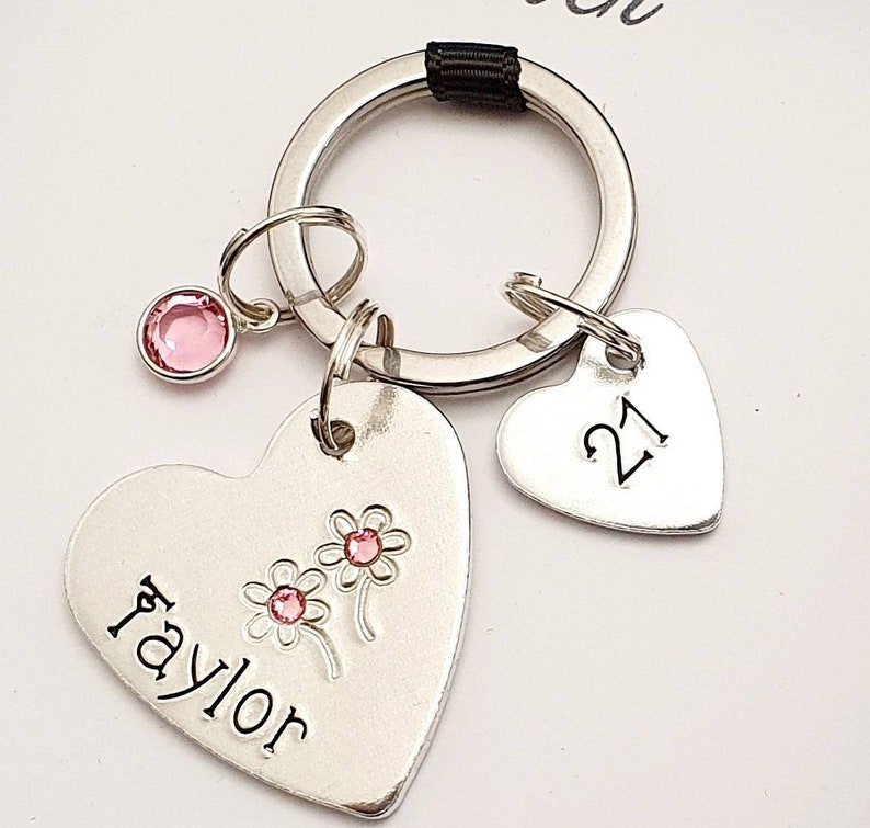 May include: Silver heart-shaped keychain with the name "Taylor" engraved on it, two pink flowers, and a pink rhinestone. A smaller silver heart-shaped charm with the number "21" engraved on it is attached to the keychain.