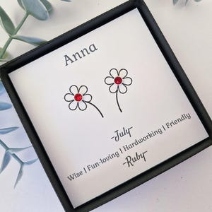 May include: A framed print with a black border shows two daisy illustrations with red gem centres. The name "Anna" is printed at the top, with "July" and "Ruby" below the flowers. The print also includes the words "Wise | Fun-loving | Hardworking | Friendly."