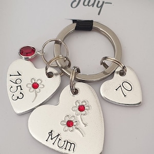 May include: Silver heart-shaped keychain with three charms. The charms are engraved with the words "Mum", "1953", and "70". Each charm has a red rhinestone flower design.