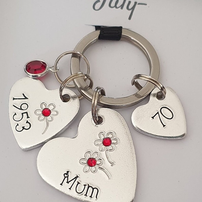 May include: Silver heart-shaped keychain with three charms. The charms are engraved with the words "Mum", "1953", and "70". Each charm has a red rhinestone flower design.