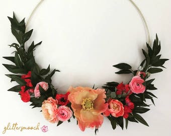 May include: A gold metal hoop wreath decorated with pink and red flowers and green leaves.