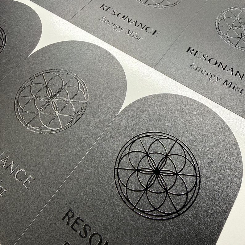 May include: A sheet of matte dark gray arch-shaped labels featuring a circular geometric pattern and the text Resonance Energy Mist embossed in a glossy finish. The labels are arranged in a grid pattern on a white backing sheet for application.